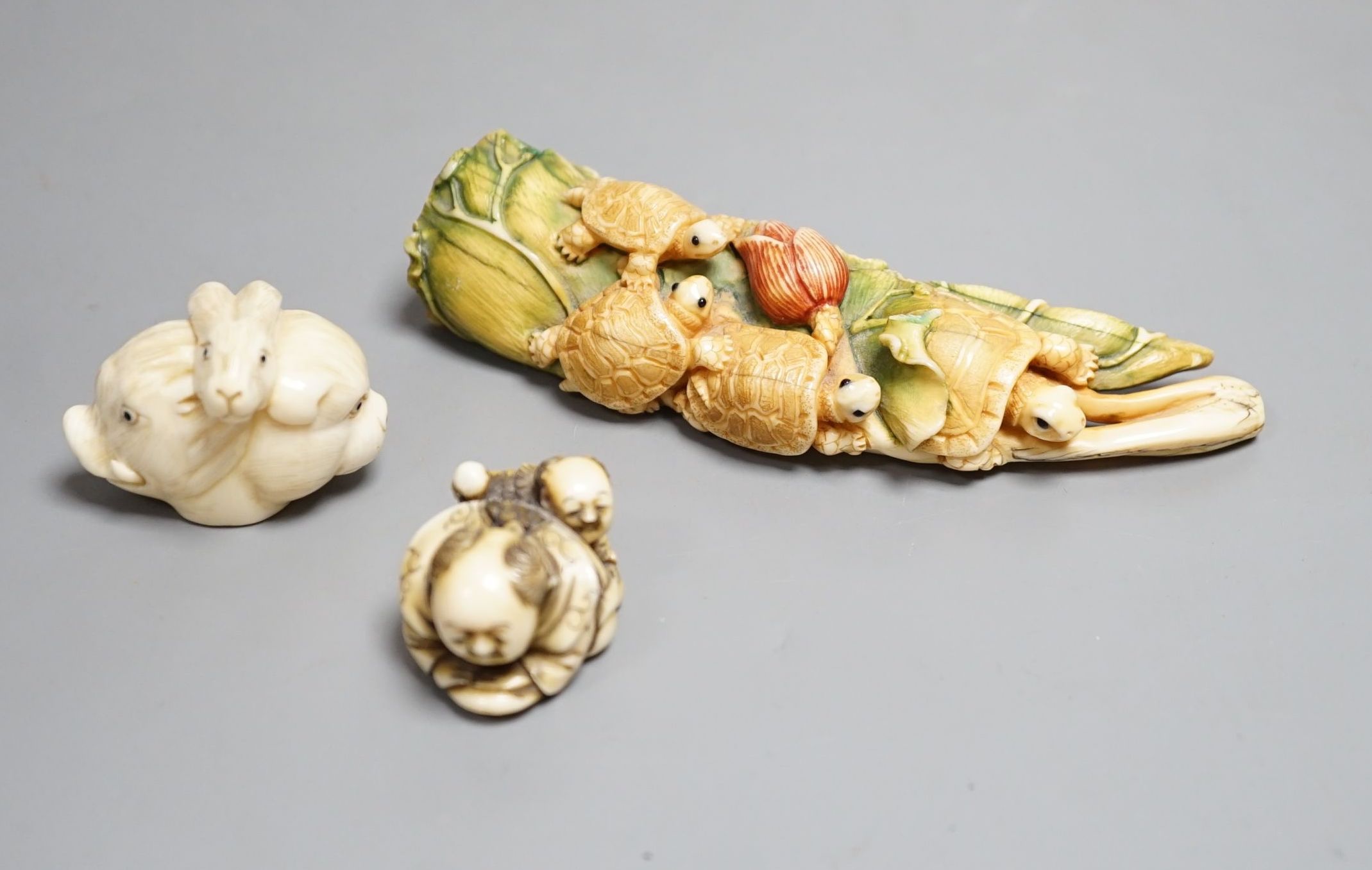 A Japanese ivory Netsuke of two boys holding a noh mask, an ivory Netsuke in the form of three animal heads and a coloured Ivory carving of tortoises on a lotus leaf, all singed (3) largest 12cm